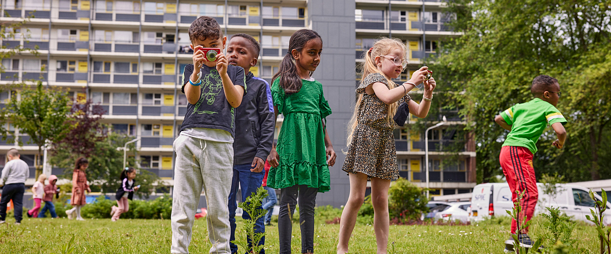 Vier jonge kinderen fotograferen planten op een grasveld voor een flatgebouw. Op de achtergrond hollen kinderen die ook samen de natuur ontdekken.