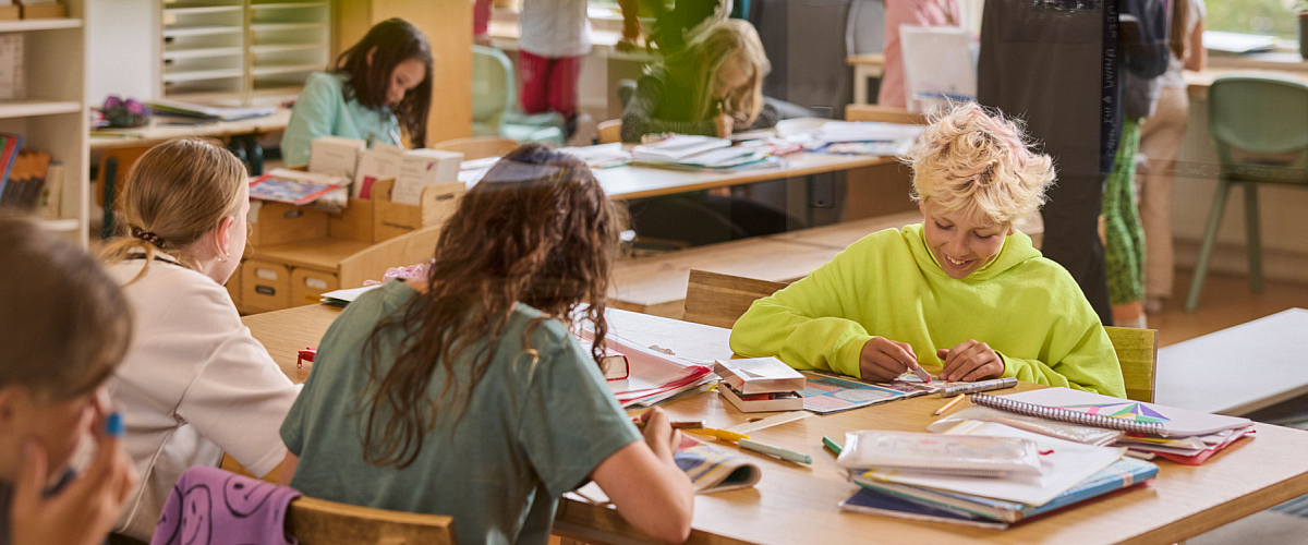 Een klaslokaal waarin kinderen in kleine groepjes zelfstandig werken aan tafels. Sommigen lezen of schrijven, anderen tekenen. De sfeer is rustig en open, met veel licht en materialen binnen handbereik. Op de achtergrond praten enkele kinderen met elkaar, terwijl vooraan een kind in een felgroene trui geconcentreerd aan een opdracht werkt.